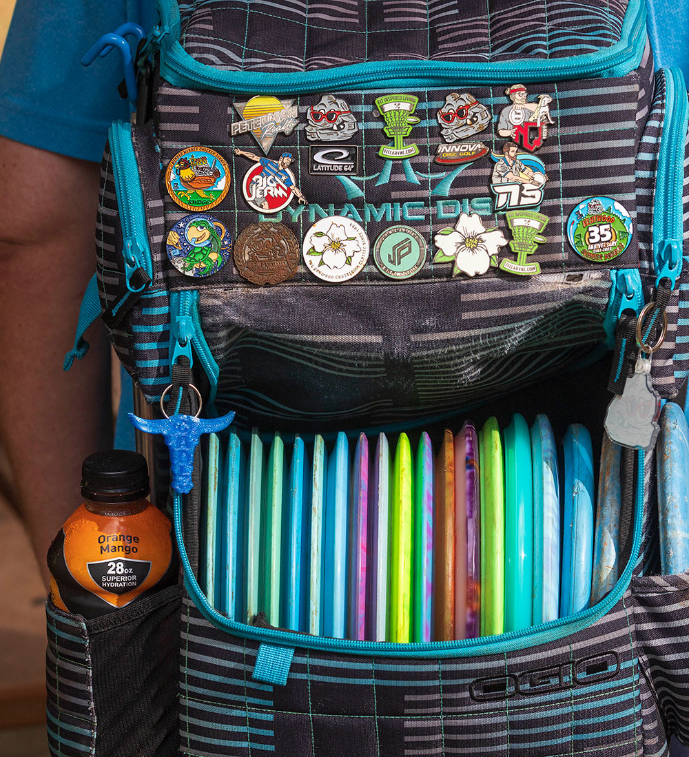 Adorned with disc golf pins, a player's backpack carries a selection of putters, mid-range discs, and drivers.