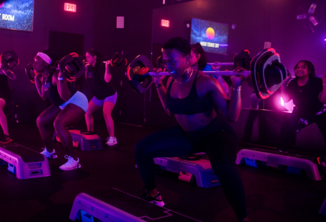 Participants standing behind step platforms squat with weight bars on their shoulders in a room lit by pink and purple lights.