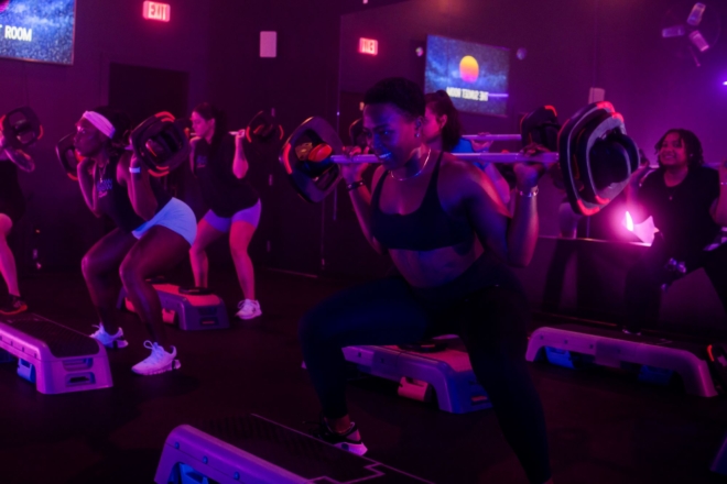 Participants standing behind step platforms squat with weight bars on their shoulders in a room lit by pink and purple lights.